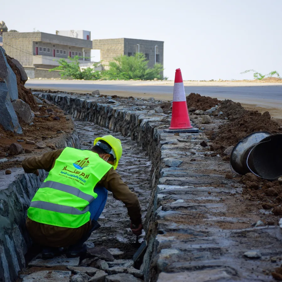 In Hajjah governorate, a mason constructs a masonry ditch channel along a hillside. The channel is designed to divert rainwater away from the road and prevent erosion, ensuring the road's stability and safety.