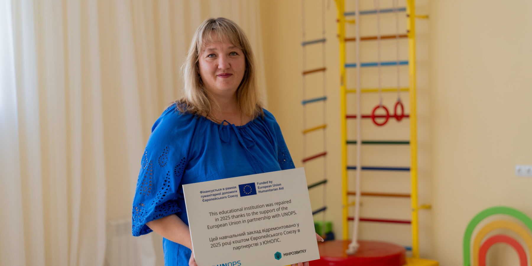 Oksana Petruk, Directora de la Institución Preescolar, sostiene un cartel en el que agradece el apoyo vital de la Unión Europea y UNOPS que ha hecho posible la renovación de la escuela.